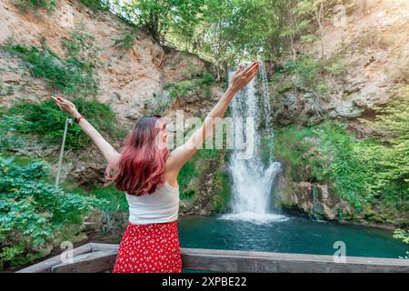 Una giovane donna gode della bellezza della cascata Veliki Buk in Serbia, in piedi nel verde lussureggiante della giungla, abbracciando la serena cascata. Foto Stock