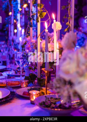 Tavolo servito per banchetti con candele e composizioni floreali. Bellissime decorazioni con luce elettrica blu e viola. Foto Stock