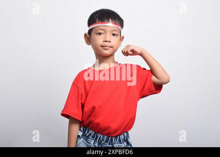 Ragazzo asiatico sicuro di sé con t-shirt rossa che flessa la forza alzando braccia e muscoli, isolato su sfondo bianco. Concetto del giorno dell'indipendenza indonesiana Foto Stock