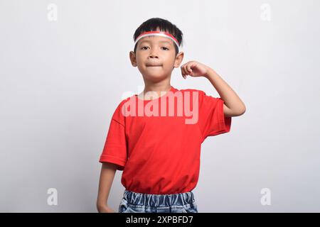 Ragazzo asiatico sicuro di sé con t-shirt rossa che flessa la forza alzando braccia e muscoli, isolato su sfondo bianco. Concetto del giorno dell'indipendenza indonesiana Foto Stock