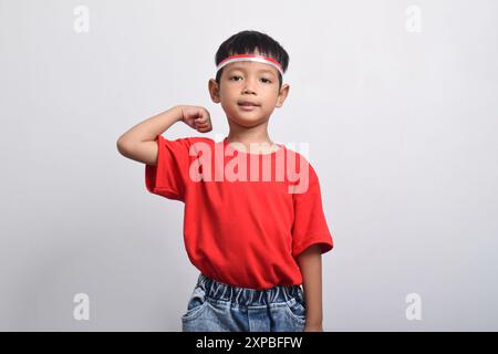 Ragazzo asiatico sicuro di sé con t-shirt rossa che flessa la forza alzando braccia e muscoli, isolato su sfondo bianco. Concetto del giorno dell'indipendenza indonesiana Foto Stock