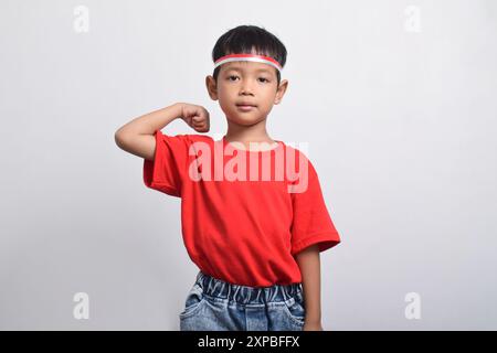 Ragazzo asiatico sicuro di sé con t-shirt rossa che flessa la forza alzando braccia e muscoli, isolato su sfondo bianco. Concetto del giorno dell'indipendenza indonesiana Foto Stock