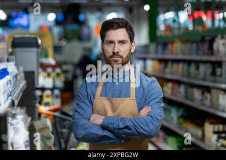 Dipendente di un negozio di fiducia in piedi in una corsia di un supermercato con le braccia incrociate. Il lavoratore che indossa il grembiule guarda la fotocamera con grande serietà. Scaffali riforniti con prodotti in background. Foto Stock