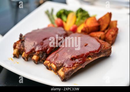 Costolette di maiale grigliate condite con speciale salsa barbecue, patatine fritte e verdure su piatto bianco nel ristorante Foto Stock