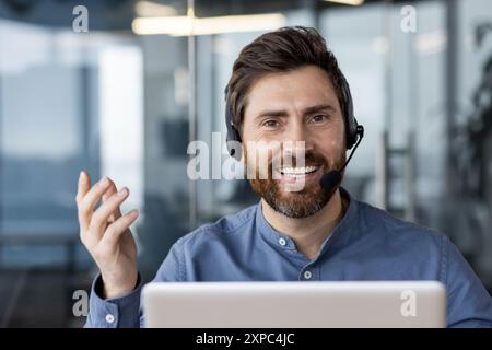 Uomo d'affari felice che indossa la cuffia sorridente durante le videochiamate in ufficio. Uomo sicuro di sé con notebook che partecipa a riunioni virtuali. Concetto di lavoro remoto, assistenza clienti e comunicazione. Foto Stock