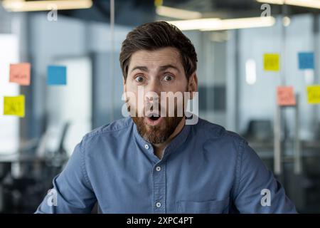 Uomo d'affari sorpreso con la barba che indossa una camicia blu in un ufficio moderno che reagisce con gli occhi larghi e la bocca aperta. Lo sfondo include pareti in vetro e note adesive colorate. Foto Stock