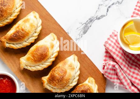 Empanadas di manzo argentino su una tavola di legno, fette di limone, salsa calda e un tovagliolo rosso e bianco su sfondo di marmo bianco. Foto Stock