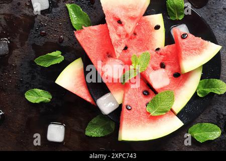 Fette di cocomero succoso maturo su un piatto nero su sfondo scuro con menta e cubetti di ghiaccio. Vista dall'alto. Un rinfrescante frutto estivo. Ingredienti Foto Stock