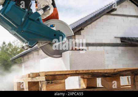 Un lavoratore utilizza un utensile elettrico per tagliare blocchi di calcestruzzo su pallet di legno in un cantiere edile sotto un cielo limpido. Foto Stock