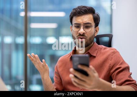 L'uomo in un ufficio moderno guarda lo smartphone con confusione, indossando occhiali e camicia arancione, facendo gesti con la mano. Indica frustrazione o malinteso Foto Stock