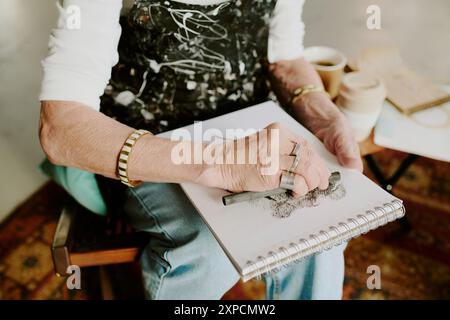 Vista ad angolo alto primo piano medio delle mani di un'artista donna anziana irriconoscibile che disegna uno schizzo con matita a carbone Foto Stock