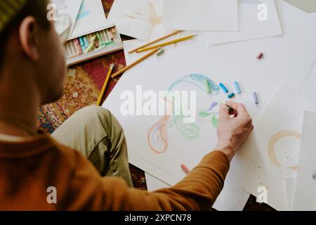Vista ad alto angolo di uno schizzo di disegno maschile non riconoscibile su un foglio di carta con bastoncini pastello Foto Stock