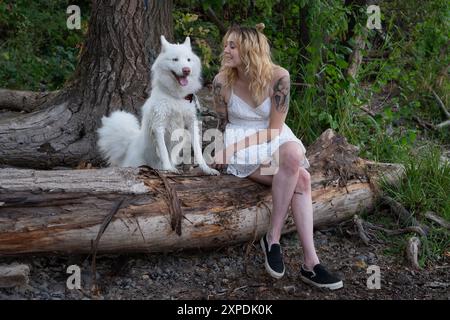 Una donna bionda con un vestito bianco si siede su un tronco con un cane bianco vicino al fiume Willamette Foto Stock