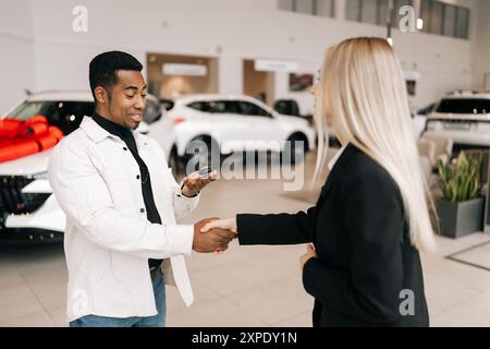 Felice e soddisfatto proprietario di auto africane che ottiene la chiave da una donna sorridente concessionaria di auto presso la concessionaria. Allegro maschio nero che prende la chiave per comprare l'auto e sigillare Foto Stock