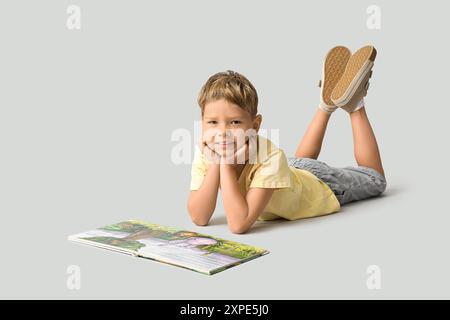 Bambino carino sdraiato sul pavimento e legga un libro su sfondo grigio Foto Stock