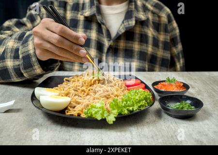 Un uomo sta mangiando un piatto di spaghetti istantanei con una forchetta e bacchette. Il piatto è ripieno di varie verdure e un uovo sodo. L'uomo è enjoyi Foto Stock