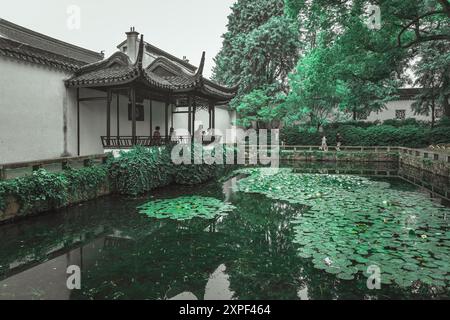 Suzhou, Cina - 11 giugno 2024: Un padiglione cinese tradizionale si trova sul bordo di uno stagno coperto da giglio, il riflesso dell'edificio visibile Foto Stock