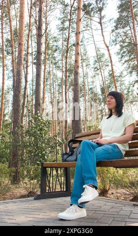 Giovane donna in abiti casual seduta sulla panchina nel parco. Ragazza in jeans blu e maglietta gialla che si rilassa nella pineta. Tranquillità nella vita Foto Stock