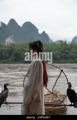 Scatto verticale di una ragazza di Hanfu su una zattera di bambù con cormorani che fissano i suoi abiti tradizionali Foto Stock