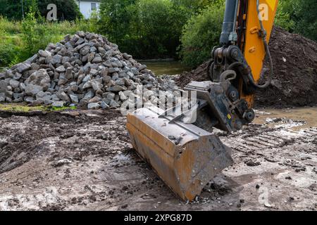 Un grande escavatore è posizionato su un cantiere, spostando lo sporco mentre è circondato da mucchi di rocce e vegetazione. Foto Stock