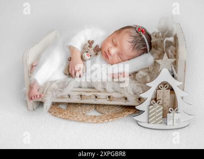 La neonata con cervo giocattolo dorme su un piccolo letto con un piccolo albero di Natale nelle vicinanze durante Una sessione fotografica presso lo Studio Foto Stock