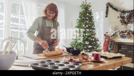 Ritratto di una donna anziana che prepara biscotti di pan di zenzero per Natale a casa. La nonna preparava felicemente dei dolci da condividere con la famiglia e gli amici, preparandosi per la festa e la cena Foto Stock