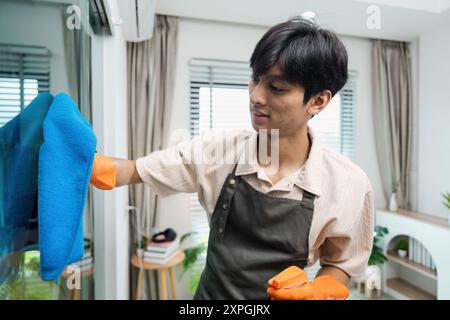 Finestra per la pulizia di Young Man con panno blu e guanti arancioni in interni moderni Foto Stock