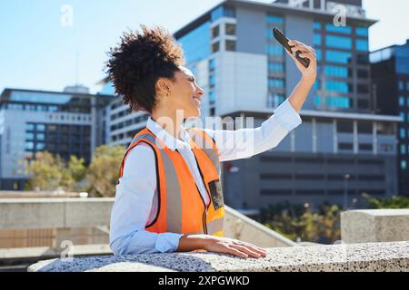 Lavoratrice edile che porta un selfie con uno smartphone in un ambiente urbano Foto Stock