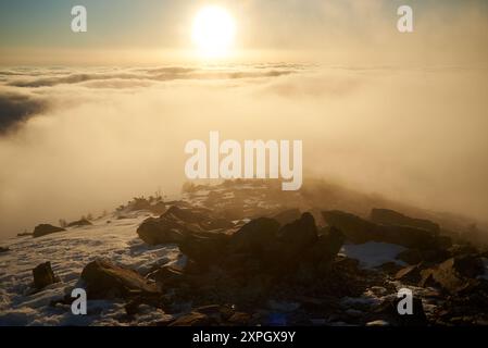 Alba sulla cima innevata della montagna. La calda luce dorata del sole che sorge bagna le nuvole in delicate sfumature di arancione e rosa. Neve e rocce sparse in primo piano aggiungono texture e profondità alla scena. Foto Stock