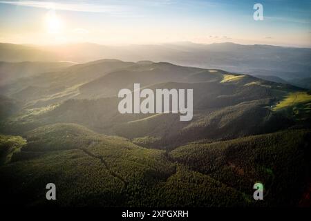 Vista aerea del tranquillo paesaggio montano al tramonto. Colline boscose immerse in una calda luce dorata, creano un'atmosfera tranquilla. Colline ondulate si estendono in lontananza, coperte da una fitta vegetazione lussureggiante. Foto Stock