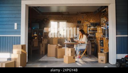Donna d'affari ispanica che usa un computer portatile in garage. Titolare di una piccola impresa che compila gli ordini dei clienti online. Prodotti in scatole di cartone pronti per la spedizione. Imprenditore che lavora a casa. Foto Stock