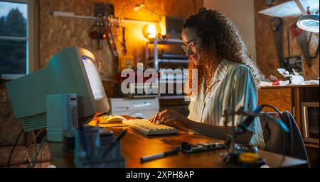 Hispanic Female Engineer Using Old Desktop computer in Nineties retro Garage la sera. Donna intelligente che scrive codice, lavora su dispositivi portatili innovativi, inizia Tech Startup Company. Foto Stock