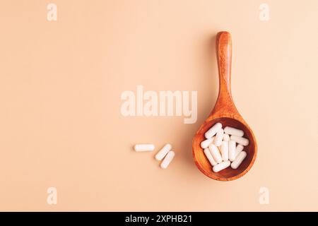 Cucchiaio di legno con capsule bianche su sfondo beige, vista dall'alto. Integratori alimentari e rimedi naturali in una composizione semplice ed elegante. Foto Stock