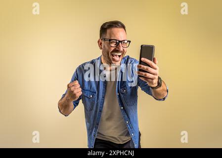 Freelance di successo che legge buone notizie sul telefono cellulare e urla allegramente su sfondo beige. Giovane vestito con camicia in denim Foto Stock