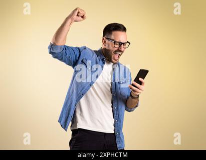 Uomo d'affari eccitato vestito con camicia in denim che urla e alza la mano mentre legge buone notizie sul cellulare. Giovane che celebra il successo mentre s Foto Stock