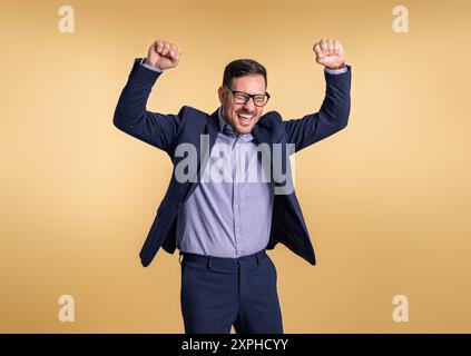 Giovane uomo d'affari di successo con gli occhi chiusi e le braccia alzate urlando allegramente isolato su sfondo giallo. Uomini eleganti e professionali danci Foto Stock