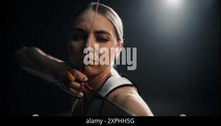 Ritratto di un bellissimo arciere femmina che regge un moderno arco composto, che mira a un bersaglio. Giovane atleta che disegna la Freccia in un concorso Stadium o in uno studio oscuro per la pubblicità online Foto Stock