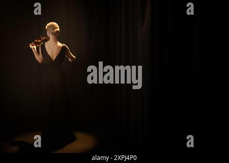Full shot posteriore di una cantante di musica classica che suona il violino indossando un abito posteriore con taglio basso in piedi da sola sotto la luce del proiettore sul palco scuro con tende nere e spazio per le copie Foto Stock