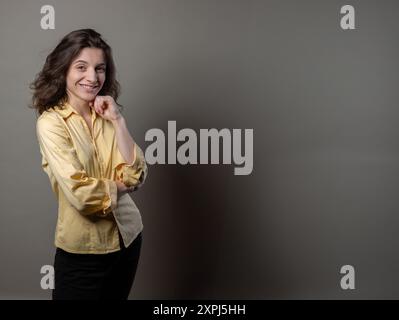 Una donna con una camicia gialla è in piedi davanti a un muro grigio. Sta guardando la telecamera con un'espressione seria Foto Stock