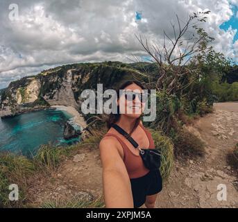 Una donna sta facendo un selfie su una spiaggia con una splendida vista sull'oceano. Sta indossando occhiali da sole e una borsa nera Foto Stock