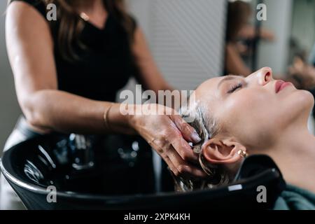 Primo piano di uno shampoo schiumoso per parrucchieri irriconoscibile sui lunghi capelli biondi di una bella cliente femminile. Primo piano del lavaggio da parrucchiere Foto Stock