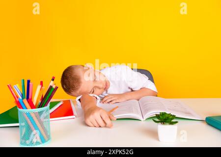 Foto di un ragazzo simpatico e assonnato seduto alla scrivania che fa i compiti a casa si addormenta isolato su uno sfondo di colore giallo Foto Stock