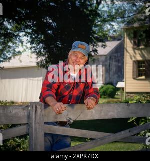 Agricoltore appoggiato alla recinzione. Camicia, berretto, cappello in flanella. Foto Stock