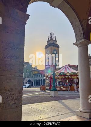 Il paesaggio della Citta bassa con il cielo del tramonto, la Torre commemorativa e la giostra in stile vintage in Piazza Vittorio Veneto attraverso l'antico arco in pietra, Bergam Foto Stock