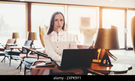 Una donna d'affari con una camicia bianca siede a un tavolo con un notebook in un ufficio moderno e ben illuminato. Lo sfondo presenta grandi finestre ed eleganti Foto Stock