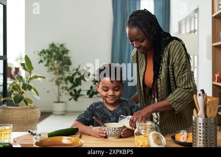 Felice giovane donna afroamericana che versa latte fresco dal biberon in una ciotola con fiocchi di mais mentre prepara la colazione per il suo bambino Foto Stock