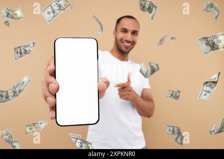 Uomo felice che punta al telefono cellulare con schermo vuoto sotto la pioggia di denaro su sfondo beige scuro, messa a fuoco selettiva Foto Stock