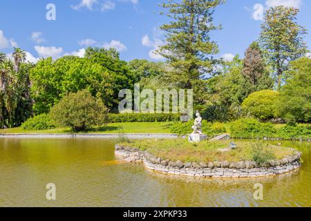 Serena scena di una statua di una donna seduta su una piccola isola in un lago, circondata da vegetazione lussureggiante e alberi Foto Stock