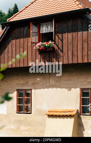 Una pittoresca casa rustica presenta una finestra aperta adornata da fiori vibranti, che mostra una calda facciata in legno. La scena cattura l'essenza della pace Foto Stock