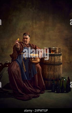 Uomo, monaco in abito d'epoca seduto accanto a botte di legno di birra, con birra e tazze di birra sopra, mangiando di nuovo patatine scure sullo sfondo vintage Foto Stock